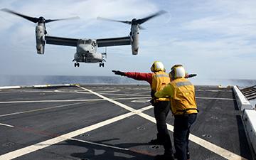 Two men wearing yellow vests helping a military plane land on a ship at sea