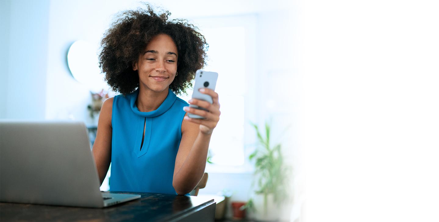 Woman in blue shirt, smiling and looking at her phone in front of a laptop, enjoying Viasat's internet packages